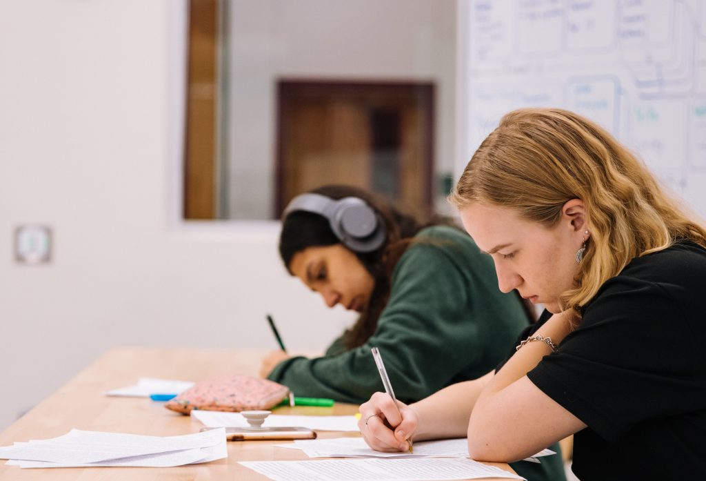 Two students focused on writing notes in a well-lit classroom, with educational materials on the table and a whiteboard with written concepts.